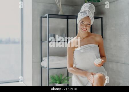 A woman with a towel on her head smiles, holding a skincare product in a minimalist spa Stock Photo