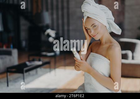 Chic woman with towel on hair, holding a lotion tube, in a stylish interior. Stock Photo
