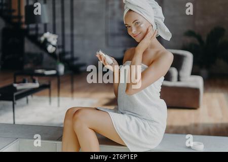 Elegant woman applying facial cream in a modern home, wrapped in a towel Stock Photo