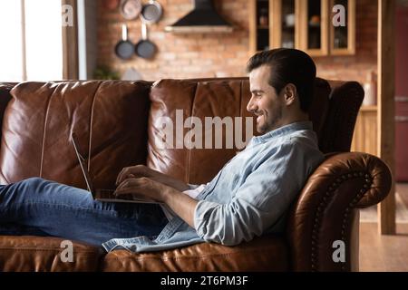 Side view smiling man using laptop, having fun, relaxing on cozy couch Stock Photo