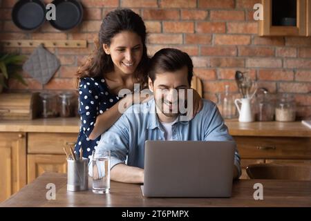 Happy young man and woman hugging, using laptop in kitchen Stock Photo