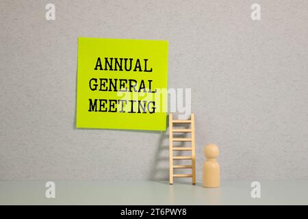 AGM Annual General Meeting is written in red on a white piece of paper on a light yellow background next to a laptop, pen, magnifying glass, glasses a Stock Photo