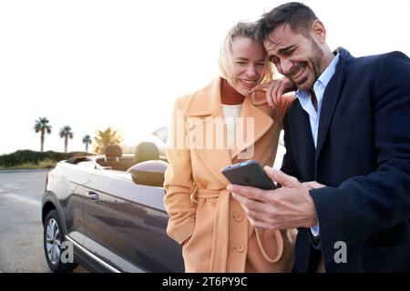 Happy mature couple using a smartphone outside the convertible car and laugh Stock Photo