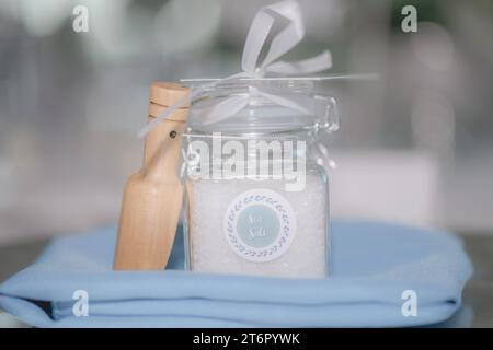 Beautiful Sea Salt filled clear jar with white bow wedding favor with wood on blue blanket in front of white bokeh Stock Photo