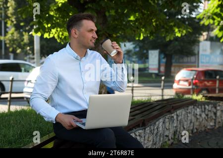 Handsome man with laptop drinking coffee on bench outdoors. Space for text Stock Photo