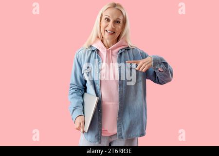Mature female programmer with laptop pointing at something on pink background Stock Photo