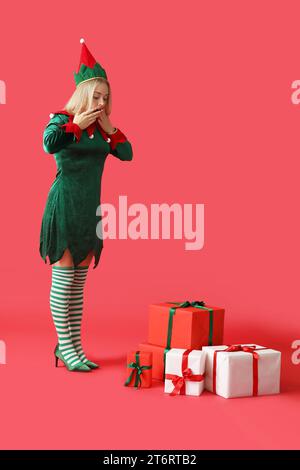 Shocked young woman in elf costume with Christmas gift boxes on red background Stock Photo