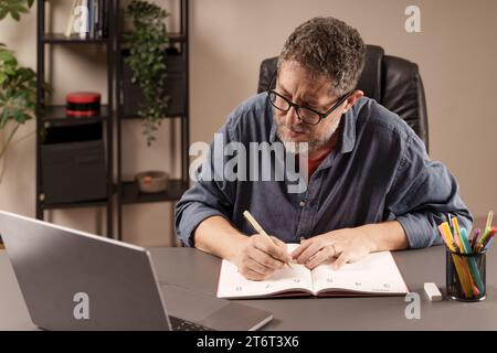 Man focused on writing in his planner, organizing tasks while working on his laptop in a home office setting. Stock Photo