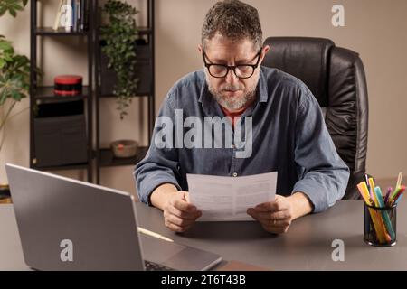 Concentrated mature man reading a paper document at his home office desk, engaged in work-related research or review - professional reviewing a paperw Stock Photo