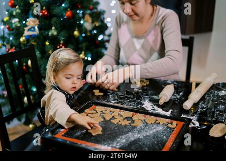 Mom makes Christmas cookies with cutouts, and a little girl puts them on a baking sheet Stock Photo