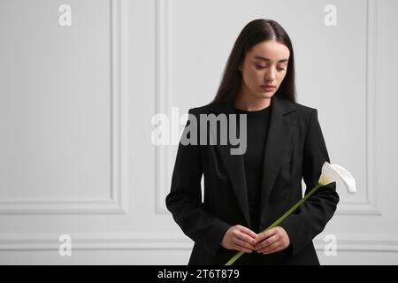 Sad woman with calla lily flower near white wall, space for text. Funeral ceremony Stock Photo