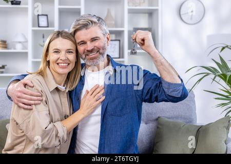 A couple of affectionate senior people in love are sitting on the sofa in their own living room and holding the key to a new home, moving to a new apartment, buying their own house. Stock Photo