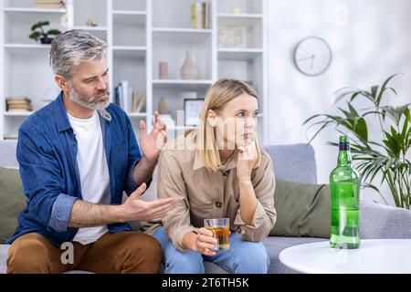 Upset couple sitting in living room on sofa, alcoholic woman drinking a lot of strong alcohol alone, man trying to stop her, discuss everything, relationship problems, alcohol addiction. Stock Photo