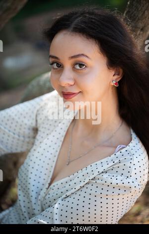 A beautiful young woman enjoying the warm summer day, leaning against a tree in a lush green outdoor setting Stock Photo