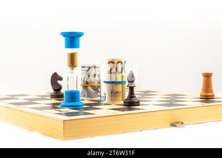 Front angle view on chess board with selected black and white chess figures, and hourglass and rolled euro bank notes and American dollar bills. Conce Stock Photo