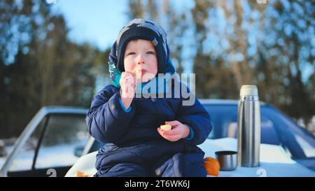 The boy eats tangerines in winter sits on the car. Stock Photo