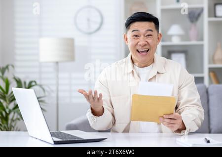 Surprised Asian man sitting at desk in home office, holding open envelope in hands, can't believe his luck. Korean senior man is happy with the win, good news, look at the camera. Stock Photo