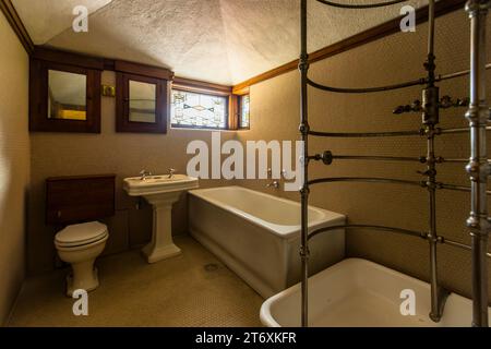 The Frederick C. Robie House houses, on its top floor in Chicago, Illinois, United States, a bathroom with a bathtub set into mosaic tiling and a separate shower basin with an elaborate needle shower. A spiral-coiled pipe frame with numerous nozzles encloses the shower basin on the right of the frame, while a wall-mounted toilet and a pedestal washbasin with two mirrored wall cabinets stand on the left. A small leaded-glass window with a diamond pattern lights the room from above. Since 2019, the house has been one of eight of Wright's buildings on the UNESCO World Heritage List. Stock Photo