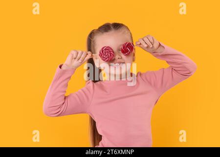 Happy little girl with bright lollipops covering eyes on orange background Stock Photo
