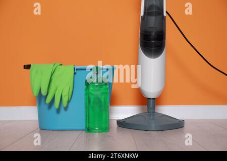 Modern steam mop, bucket with gloves and bottle of cleaning product on floor near orange wall Stock Photo