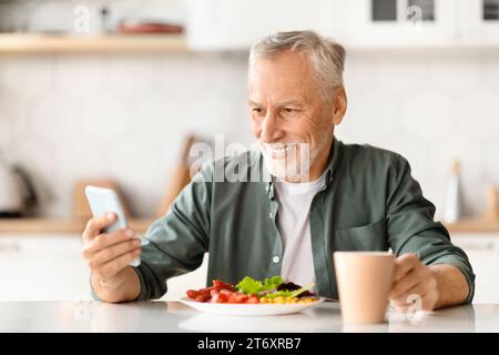 Happy senior man using smartphone while having breakfast at kitchen Stock Photo