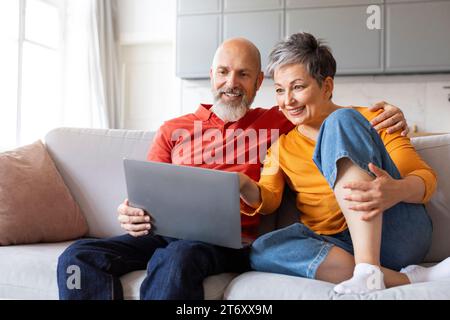 Smiling Senior Spouses Resting With Laptop In Living Room Interior Stock Photo