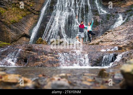 Young couple practicing yoga near the beautiful waterfall, standing in a tree position. Nature, relaxation, and partner yoga concepts. Stock Photo