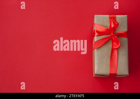 One rectangular gift box, packed in craft paper, tied with red ribbon with bow. Lies on the right on red background. View from above. Flat lay. Stock Photo