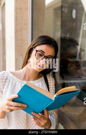 Young lady with glasses reading book near window Stock Photo