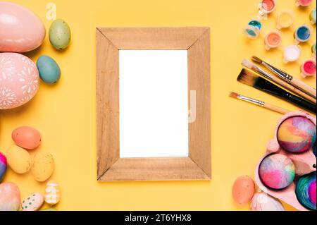 Wooden blank white frame with easter eggs paint brushes watercolor paint yellow background Stock Photo