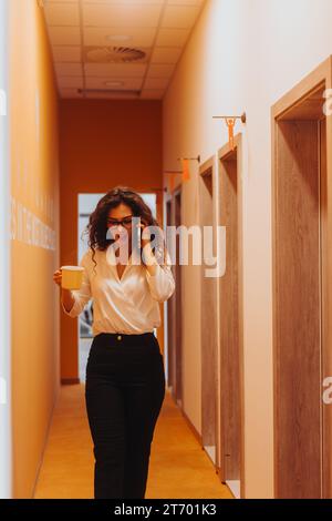 A modern, successful woman negotiates a project, enjoying tea while talking on the phone in a positive corporate office environment. Stock Photo