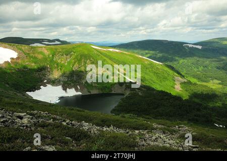 A small lake in an intermountain basin with remains of snow on the slopes under a cloudy summer sky. Ivanovskie lakes, Khakassia, Siberia, Russia. Stock Photo
