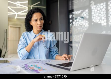 A focused thoughtful businesswoman sits in a modern office at a table in front of a laptop, holds glasses in her hands and looks intently at the computer screen, errors in reports. Stock Photo