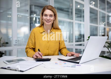 Smiling female boss working in bright office at table in front of laptop, counting on calculator, happy blonde smiling and looking at camera. Stock Photo