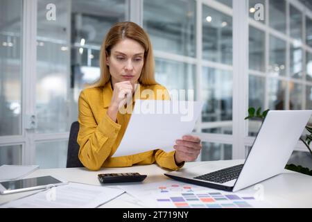 Upset blonde businesswoman working in a modern office, sitting at a desk in front of a notebook, thoughtfully working with business papers, carefully reviewing important information. Stock Photo