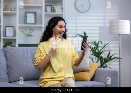 A young curly Indian woman sits on a sofa in a bright living room, holds a phone in her hand, speaks on a video call with relatives and friends, greets, smiles and waves her hand at the screen. Stock Photo