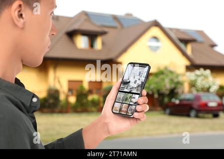 Man using smart home security system on smartphone near house outdoors. Device showing different rooms through cameras Stock Photo