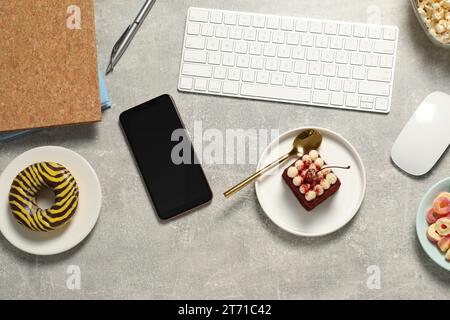 Bad eating habits at workplace. Keyboard, smartphone and different snacks on light grey table, flat lay Stock Photo