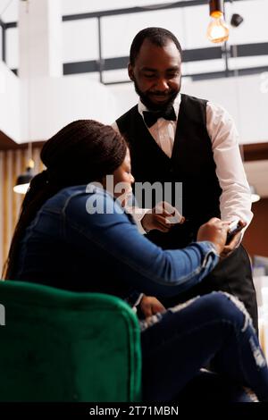 African American woman guest making contactless payment at hotel while resting in lobby. Smiling friendly waiter using NFC enabled card reader to let customer pay for order in restaurant or cafe Stock Photo