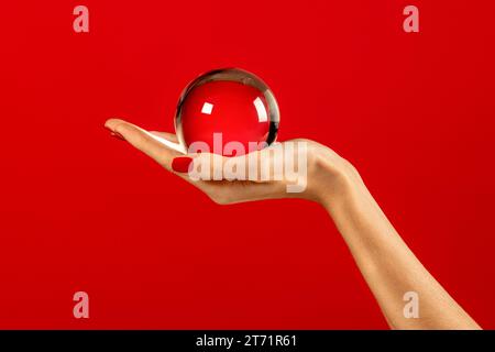 Anonymous slim young female with nails polished fingers holding transparent and reflecting glass sphere in hand against red background in light Stock Photo