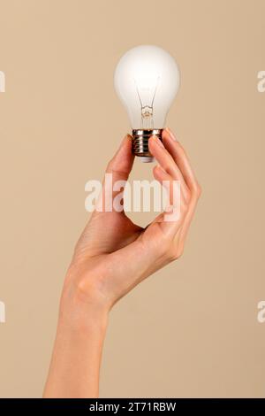 Anonymous young slim female with slender fingers holding aloft light bulb with filament in display in hand against brown background Stock Photo