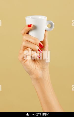 Anonymous young slim female hand with polished nails of slender fingers holding aloft coffee cup in light against brown background Stock Photo