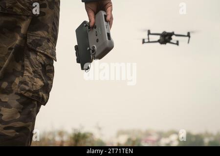 a man in a military uniform with a UAV control panel in his hands, a drone flying in the background in nature Stock Photo