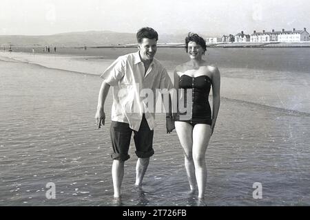 1960s, historical, a young adult couple standing in seawater beside a beach holding hands, England, UK. The young woman is in a swimsuit, while the young mn has his trousers rolled up, a traditional male thing to do at the seaside. Stock Photo
