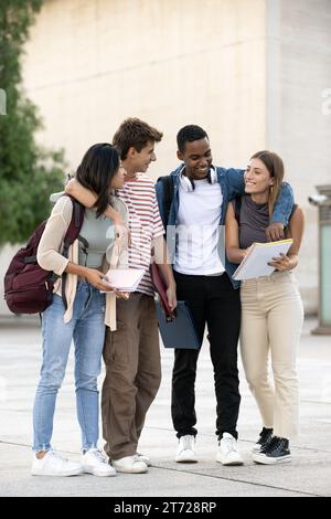 Young diverse friends walking together in embrace while reading notebooks. Joyful multiracial group of students walking hugging each other and Stock Photo