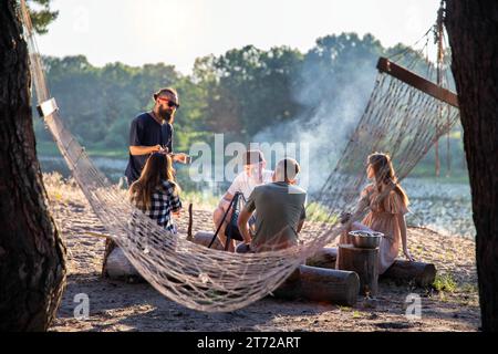 A stretched hammock on the trees against the background of a company of friends vacationing in nature in the summer on the banks of the river, campfir Stock Photo