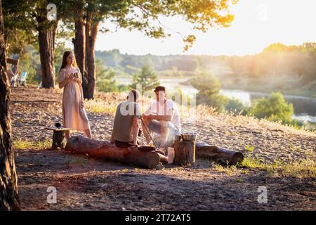 A company of friends of girls and guys have a rest in the summer in a picturesque place in the forest on the banks of the river against the backdrop o Stock Photo