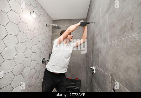 Side view of male worker using electric drilling machine while working on bathroom renovation. Man drilling wall with cordless power drill while installing shower system at home. Stock Photo