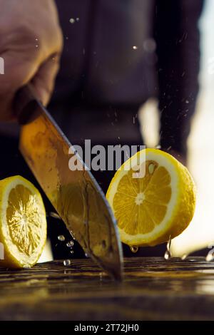 Adult man cutting lemon in half with knife. Human with knife in hand separate into two pieces juicy and sour fruit that lie on table. Yellow fruitage Stock Photo