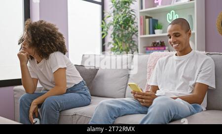 Beautiful couple sitting at home, unhappy on a roomy sofa while confident boyfriend happily engages with smartphone, problem stirring up relationship Stock Photo